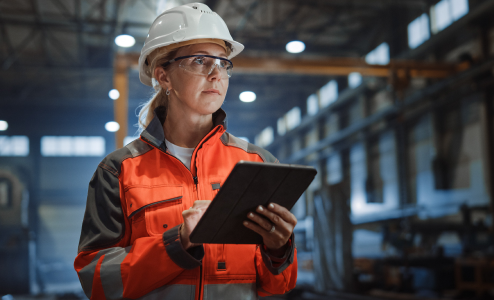 Female engineer in hard hat and safety glasses holds tablet computer in a large industrial factory, reviewing production data for manufacturing operations management. Orange and gray workwear visible.
